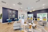 Located at 4659 Dawson Blvd in Atlanta, GA, this single-floor community amenity space offers a thoughtfully designed environment that blends social, productive, and collaborative functions. The main lounge area is anchored by a striking striped accent wall with a wall-mounted flat-screen TV, complemented by deep navy blue walls that contrast sharply with crisp white ceilings and light wood-toned flooring. Recessed lighting and stylish pendant fixtures with gold accents illuminate the space warmly throughout. Comfortable gray sofas and lounge chairs are arranged around geometric-patterned area rugs and marble-topped coffee tables, creating an inviting gathering atmosphere. A dedicated workstation zone features a white built-in desk with vibrant yellow chairs, fully equipped with computer monitors, ideal for remote work or study. Large windows and glass doors flood the interior with natural light and provide views of the surrounding greenery and outdoor deck. A separate conference room offers a glass-top table surrounded by bold yellow velvet chairs, accented by contemporary abstract artwork and a dramatic geometric chandelier, with a large window framing outdoor views. Curated artwork and lush indoor plants complete the polished, upbeat aesthetic of this well-appointed community space.