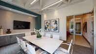 Located at 2429 Americana Blvd in Orlando, Florida, this single-floor professional space is designed to support productivity and collaboration in a polished, contemporary setting. The centerpiece of the space is a well-appointed conference room featuring soaring ceilings with exposed white structural beams and integrated linear lighting that creates a bright, focused atmosphere. A large white rectangular conference table is surrounded by sleek white upholstered chairs, accommodating a comfortable number of attendees. A wall-mounted flat-screen display is positioned above a long, built-in wood-grain credenza with ample cabinet storage, ideal for presentations and media use. Deep teal accent walls add a bold, sophisticated contrast to the neutral palette throughout. Large windows with draping curtains invite natural light and provide a view of the outdoors, enhancing the room's open feel. Curated abstract artwork adds a creative touch to the walls. Adjacent to the conference room, a dedicated business center workstation features a floating white countertop, a computer, a multifunction printer, and vibrant orange accent chairs beneath geometric pendant lighting against a dramatic dark patterned feature wall. Glass-paneled doors connect the areas, maintaining visual openness while preserving acoustic separation.