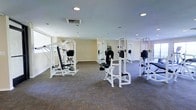This single-floor fitness center offers a clean, functional environment designed to support a comprehensive workout routine. The space is outfitted with multiple white-framed strength-training machines, including cable systems, adjustable benches, and resistance stations, all arranged with ample spacing to allow for comfortable, unrestricted movement between equipment. The room features neutral beige walls and plush carpeted flooring in a subtle multi-tone pattern, contributing to a welcoming and practical atmosphere. Recessed ceiling lights provide consistent, even illumination throughout the space, while a large window along one wall invites natural light, enhancing the overall brightness and openness of the room. The high ceiling adds to the sense of spaciousness, making the facility feel airy and unconfined. A decorative potted plant near the center of the room adds a touch of greenery, softening the otherwise utilitarian aesthetic. The layout is well-organized, maximizing the usable floor area while maintaining clear pathways between machines. Overall, this fitness center strikes a balance between functionality and comfort, making it a practical and inviting space for strength training and general fitness activities.