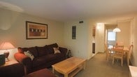 This single-floor residence offers a welcoming and functional layout designed for comfortable everyday living. The living room serves as the central gathering space, furnished with a plush, dark-toned sofa and a natural wood coffee table that grounds the room with warmth. Neutral, light-colored walls and carpeted flooring create a soft, understated ambiance throughout. Decorative wall art adds visual interest and a personal touch to the space. A ceiling vent indicates a central air system, contributing to year-round comfort. The living area opens into a hallway that connects to additional rooms, including what appears to be a kitchen or dining area visible in the background, featuring cabinetry and tiled surfaces. The corridor layout allows for easy flow between spaces, making the residence feel organized and accessible. The overall design aesthetic is simple and practical, with warm lighting enhancing the inviting atmosphere. This space is well-suited for those seeking a straightforward, comfortable home with a cohesive interior palette and a layout that balances openness with defined living zones.
