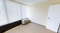This single-floor space presents a clean, straightforward layout with neutral finishes throughout. The room features wall-to-wall carpeting in a beige tone, contributing to a quiet and functional atmosphere. A textured popcorn ceiling adds a classic touch overhead, while smooth, off-white walls provide a blank canvas adaptable to various uses.

A through-wall PTAC (packaged terminal air conditioner) unit is installed at the base of the exterior wall, offering self-contained climate control directly within the room. A large window fitted with full-length vertical blinds spans a significant portion of one wall, allowing for adjustable natural light while maintaining privacy. The blinds appear to permit generous daylight when open, brightening the interior.

The room's corner configuration, with two adjoining walls meeting at a right angle, creates a sense of defined space. Minimal baseboards line the perimeter of the room. Overall, the space is unfurnished and move-in ready, offering a neutral, functional environment suitable for residential or light office use.