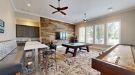 Located at 85 Washington St in Plainville, MA, this single-floor game room is a thoughtfully designed entertainment space that blends rustic charm with modern amenities. The main room is generously sized and anchored by a full-size pool table with a vibrant blue felt surface, complemented by a classic wooden shuffleboard table positioned nearby. A striking reclaimed wood accent wall serves as a focal point, housing a large flat-screen TV and adding warm, textural character to the space. An arcade cabinet adds a nostalgic touch, rounding out the entertainment options. The room is finished with elegant crown molding, a ceiling fan for comfort, and a bold patterned area rug that ties the design elements together. Large windows along one wall allow abundant natural light to fill the space while offering views of the surrounding greenery. A bar-height seating area with stylish stools provides a comfortable spot for spectators. The entryway hallway features light hardwood-style flooring, creating a clean transition into the main game area. The overall ambiance is lively, welcoming, and well-suited for social gatherings and recreation.