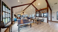 This single-floor community amenity space blends rustic charm with modern design through a striking architectural framework of soaring vaulted ceilings and prominent exposed dark wood beams. Light hardwood flooring flows throughout, tying together the distinct functional zones seamlessly.

The central gathering area serves as the heart of the space, featuring a full-size ping pong table and a substantial shuffleboard table, ideal for recreational use. Decorative geometric lattice panels add an artistic focal point, while elegant orb and lantern-style pendant chandeliers cast warm, inviting light throughout.

Adjacent to the game area, a well-equipped kitchenette offers a large island with bar seating, navy blue cabinetry with stainless steel appliances, shiplap-accented walls, and white quartz countertops. Oversized lantern pendants hang above the island, reinforcing the space's warm industrial-farmhouse aesthetic.

Multiple lounge zones provide comfortable seating, including plush sectional sofas, leather accent chairs, and a large wall-mounted TV for entertainment. A dining area with high-top tables and modern chairs offers additional gathering options.

Black-framed grid windows line the walls throughout, flooding the interior with natural light and offering views of the surrounding outdoor amenity areas, including what appears to be a pool courtyard. The overall ambiance is welcoming, versatile, and thoughtfully designed for both social gatherings and relaxed everyday use.