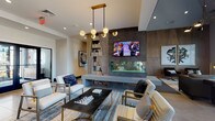 This expansive single-floor space is thoughtfully designed to accommodate a variety of social and recreational activities, with distinct zones that flow seamlessly into one another. The centerpiece of the main lounge area is a striking concrete-clad feature wall housing a linear see-through fireplace, wall-mounted television, and built-in bookshelf, anchored by a sculptural multi-globe brass pendant light. Comfortable seating arrangements with gold-accented chairs surround a dark marble coffee table, all set atop layered area rugs over large-format tile flooring.

A secondary lounge area features an oversized sectional sofa positioned in front of expansive gridded windows that invite generous natural light and exterior views. Adjacent to this, floor-to-ceiling sliding glass doors open to an outdoor patio area with greenery and seating visible beyond.

The dining area offers soaring ceilings, large windows, and upholstered chairs arranged around wood-topped tables beneath a bold multi-arm chandelier. A games room with a billiards table is visible beyond. The kitchenette presents light wood cabinetry with black hardware, dark countertops, a stainless undermount sink, under-counter appliances, open metal shelving, and a geometric hexagonal tile backsplash.