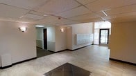 This single-floor lobby serves as the primary entry point for the building, offering a functional and organized common area. The space features a combination of large light-toned tiles near the entrance and darker tile accents at the threshold, creating a defined transition as you step inside. A suspended drop ceiling with acoustic tiles spans the area, keeping the layout clean and uniform overhead.

A wall-mounted sconce provides warm accent lighting along the interior corridor, complementing the recessed ceiling lights that illuminate the space evenly. The walls are finished in a neutral beige tone, contributing to a calm and understated atmosphere throughout.

A prominent bank of individual mailboxes lines one wall, indicating the building's multi-unit nature and serving as a central convenience for residents. The corridor leading away from the main lobby area features dark carpeting and leads toward what appears to be a door with access-controlled entry, suggesting additional secured areas beyond.

The glass entrance doors at the far right allow natural light to filter into the lobby, offering a view of the outdoors and creating an inviting atmosphere upon arrival. Overall, the lobby presents a practical, well-organized space designed to meet the everyday needs of building occupants.