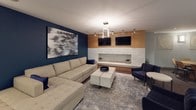 This single-floor amenity lounge is thoughtfully designed to offer a variety of social and recreational experiences within one open, well-appointed space. The layout is divided into several distinct functional zones that flow seamlessly into one another.

At the heart of the lounge is a comfortable seating area anchored by a large sectional sofa in neutral tones, set against a bold deep-blue accent wall adorned with striking large-scale abstract artwork. A linear fireplace with a textured white stone surround serves as a focal point, flanked by warm wood cabinetry and dual wall-mounted flat-screen televisions overhead. A geometric pendant light fixture adds a modern architectural touch to this gathering area.

Adjacent to the lounge seating is a billiards table, providing a dedicated recreation zone with its own pendant lighting and wall sconces. A round wood dining table with cushioned chairs offers a casual dining or meeting area nearby, also accented with curated artwork.

The kitchenette features light wood cabinetry, granite countertops, a mosaic tile backsplash, and an island with bar seating — ideal for entertaining. A separate conference-style dining table with a herringbone wood surface provides additional seating and a more intimate gathering spot.

Throughout, the space is finished with plush dark carpeting, recessed lighting, and a cohesive contemporary aesthetic that balances comfort with sophisticated design.