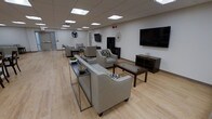 This single-floor space functions as a well-appointed communal lounge, thoughtfully designed to accommodate group gatherings and casual relaxation. The room features light-toned hardwood flooring that runs throughout, contributing to a clean, open aesthetic. Overhead, a drop-tile ceiling hosts a grid of recessed fluorescent panel lights, providing bright, even illumination across the entire space.

The seating arrangement is organized into multiple distinct zones. A large gray sofa anchors the central area, paired with a dark wood coffee table featuring decorative tile inlays. Complementing the sofa are several gray upholstered armchairs, some with patterned accent pillows, arranged to encourage conversation. A slim, industrial-style console table with metal legs sits behind the sofa, decorated with small sculptural accents.

Two wall-mounted flat-screen televisions serve different areas of the room, enhancing the space's versatility. A dark wood media console provides additional storage near one of the screens. Decorative wall art adds subtle visual interest to the neutral white walls. The overall ambiance is modern and functional, balancing comfort with a clean, contemporary design aesthetic suited for shared use.