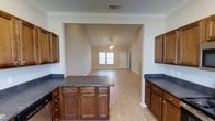 Located at 28013 Hwy 181 in Daphne, Alabama, this two-story property offers a well-designed interior that emphasizes open, connected living spaces. The kitchen is a central highlight, featuring rich, warm-toned wood cabinetry with brushed nickel hardware and dark countertops that provide a striking contrast. A kitchen island with matching cabinetry below offers additional workspace and storage, making the kitchen both functional and visually cohesive. Crown molding lines the upper perimeter, adding a refined architectural detail throughout.

The kitchen opens directly into a generous living area, creating a seamless flow ideal for everyday living and entertaining. The living space is finished with light hardwood-style flooring that extends throughout, lending warmth and continuity to the interior. Vaulted ceilings in the living area enhance the sense of openness, while a ceiling fan provides practical comfort. Natural light filters in through a front-facing window, brightening the space and connecting it to the outdoors. Neutral wall tones throughout keep the interior feeling clean and versatile. The two-story layout suggests additional living or sleeping space on the upper level, rounding out a well-structured and functional home.