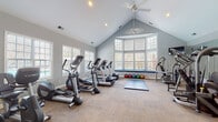 Located at 1400 Laurel Springs Dr in Durham, NC, this fitness center is a thoughtfully designed, single-floor facility built to support a comprehensive workout experience. The space is defined by its striking vaulted ceilings, which lend an open, airy quality throughout, while ceiling fans help maintain a comfortable environment during intense sessions.

Natural light pours in through an abundance of large multi-pane windows lining the walls, including a prominent bay window area fitted with a built-in padded bench — ideal for stretching or storing equipment such as the colorful medicine ball collection housed beneath it. The windows offer pleasant outdoor views, further enhancing the bright and motivating atmosphere.

The fitness floor is covered in durable carpet tile and is divided into functional zones. The cardio area is well-stocked with multiple treadmills, elliptical trainers, stationary bikes, and a rowing machine. The strength training zone features a robust selection of cable machines, multi-function weight stations, and a dedicated free weights area with a rack and adjustable bench. Full-length wall mirrors expand the sense of space and allow users to monitor their form during exercises.

Glass-paned French doors provide access to the space, and wall-mounted TVs add an entertainment element to cardio workouts. The overall ambiance is clean, modern, and functional — a well-rounded fitness facility suited for a variety of training styles.