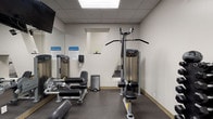 This fitness center is a dedicated exercise space designed to support a comprehensive workout routine. The room features durable rubber flooring throughout, providing both safety and comfort during training sessions. A suspended drop-panel ceiling with recessed lighting ensures the space is well-lit, while a wall-mounted mini-split air conditioning unit maintains a comfortable temperature year-round.

Large mirrors line multiple walls, creating a sense of spaciousness and allowing users to monitor their form during exercise. The equipment selection is varied and includes a multi-function cable machine suitable for lat pulldowns and cable rows, an adjustable weight bench, a leg curl and extension machine, and a dumbbell rack stocked with a full range of weights. Resistance bands are also available for additional training versatility. A wall-mounted flat-screen television is positioned for visibility throughout the room, enhancing the workout experience.

Posted fitness center rules and emergency contact information are displayed on the walls, reflecting a focus on user safety and facility management. The space also includes a compact storage area visible through a pass-through window, adding to its functional layout. The overall ambiance is clean, practical, and purpose-built for effective exercise.