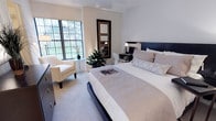 This single-floor residence offers a refined living environment highlighted by a carefully designed bedroom that serves as its centerpiece. The bedroom features a neutral color palette of soft whites, beiges, and grays, creating a calm and sophisticated ambiance throughout. A king-sized bed with a sleek dark headboard anchors the room, complemented by layered, textured bedding and decorative pillows that add depth and warmth to the space. A dedicated workspace with a dark wood desk and chair provides a functional area for productivity without compromising the room's overall aesthetic. A tall, framed mirror grouping mounted on the wall adds visual interest and reflects light effectively throughout the room. One of the standout features of the bedroom is the large multi-pane window that floods the space with natural light and frames a lush outdoor garden view, enhancing the connection between the interior and the surrounding greenery. A cozy armchair positioned near the window creates an inviting reading nook, accompanied by a small accent table and a vibrant potted plant that bring a touch of nature indoors. Soft carpeting underfoot adds to the overall comfort of the space, while recessed and accent lighting ensure a warm, well-lit environment at any time of day.