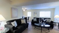 This single-story space showcases an open-concept living and dining area united by a cohesive black-and-white design aesthetic. The living area is anchored by plush black sofas adorned with patterned accent pillows, arranged around a sleek glass-topped oval coffee table that adds a contemporary touch. A ceiling fan overhead provides both function and visual interest, while multiple windows along the walls invite ample natural light, keeping the space feeling bright and airy despite the bold furnishings. The neutral-toned carpet grounds the room with warmth and comfort underfoot. A decorative accent wall featuring a geometric pattern serves as a focal point, visually separating the living and dining zones while maintaining an open, connected feel. The dining area, visible in the background, continues the monochromatic palette with dark dining chairs. Wall art and decorative accessories, including sculptural pieces on the coffee table, lend a curated, intentional quality to the overall design. The layout flows naturally between the two areas, making the space well-suited for both relaxed everyday living and entertaining.
