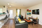 This single-story space is designed for comfortable, everyday living, with hardwood-style flooring running throughout and a cohesive, casual aesthetic accented by pops of color. The living area serves as the centerpiece of the space, offering ample room for seating with a dark leather sofa and armchair arrangement centered around a wall-mounted flat-screen television. Colorful abstract wall art adds a vibrant touch, while a glass-paneled door allows natural light to flow in alongside curtained windows. A coffee table and media console provide practical storage and surface space, completing the functional layout.

The kitchen is compact yet efficient, featuring white cabinetry, a black electric range, and light countertops. A large window above the counter looks out to greenery, filling the space with natural light and providing a pleasant backdrop during meal preparation.

The bathroom is well-proportioned, offering an extended vanity with under-sink cabinetry and a wide mirror above. Hardwood-style flooring continues into this room, maintaining visual continuity throughout the unit. Decorative wall art and coordinated accessories give the bathroom a personalized, polished feel.