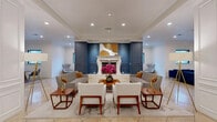 Located at 3500 Fairmount St in Dallas, Texas, this sophisticated single-floor communal space offers a thoughtfully designed open-concept layout that seamlessly blends comfort and elegance. The focal point of the main lounge area is a striking white marble fireplace set against a bold navy blue wainscoted accent wall, above which a gold-and-white abstract artwork adds a refined artistic touch. Recessed lighting and crown molding throughout lend a polished, upscale ambiance to the space.

The seating area is arranged symmetrically around the fireplace, featuring neutral-toned sofas and upholstered armchairs accented with warm wood side tables — ideal for gathering and conversation. Flooring throughout is composed of large-format cream marble tiles, contributing to the light and airy feel of the space.

Adjacent zones include a secondary lounge area with a rich navy velvet sectional sofa and natural light from a grid-pane window, as well as a dining area furnished with a communal table and modern chairs beneath globe pendant lights. A kitchenette with cabinetry and counter space supports functional use of the space. Architectural columns with wainscot detailing divide zones while maintaining visual openness. A cozy private nook with navy upholstered banquette seating completes the layout, providing an intimate retreat within the broader space.