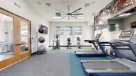 This fitness center is a thoughtfully designed, single-floor facility that provides a well-rounded exercise environment. The space is organized across three distinct zones, each dedicated to a different aspect of physical training.

The main cardio area is anchored by a row of modern treadmills positioned in front of a bold, full-wall fitness mural that displays dynamic workout imagery, creating a motivating atmosphere. A wall-mounted flat-screen television adds an entertainment option during exercise sessions. The room features elevated ceilings equipped with a large statement ceiling fan and recessed LED lighting panels, contributing to excellent air circulation and a bright, open feel.

A secondary area focuses on cycling and cross-training, housing upright and recumbent stationary bikes alongside an elliptical machine. Large windows fitted with roller shades line the walls, allowing ample natural light to filter in while offering views of the surrounding outdoor environment, which appears to include palm trees and neighboring architecture.

A third zone provides space for functional and strength training, featuring a cable resistance machine, a dumbbell rack, and a collection of stability balls on a dedicated stand. Full-length mirrors are mounted in this area to support proper form during workouts. Flooring throughout the facility consists of dual-tone carpet tiles in teal and grey, delineating the different workout zones. The facility is accessed through a warm wood-framed glass entry door, adding a refined architectural touch.