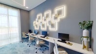 This single-floor business center is designed with a clean, contemporary aesthetic that balances functionality with thoughtful design. The main workspace features a long, continuous white desk running along one wall, accommodating multiple computer workstations equipped with monitors, keyboards, and ergonomic mesh and upholstered chairs. The seating arrangement supports both individual focus work and side-by-side collaboration.

The most distinctive design element is a gallery-style wall installation featuring overlapping square LED light fixtures with gold accents set against cool grey walls. These backlit geometric forms cast a warm ambient glow that complements the recessed ceiling lighting, creating a layered and visually dynamic atmosphere throughout the room.

A floor-to-ceiling window dressed with sheer white curtains allows natural light to fill the space, offering outdoor views and enhancing the room's openness. The flooring consists of a patterned grey carpet that adds texture underfoot and contributes to a refined, professional environment.

A framed piece of colorful artwork is displayed near the entryway, adding a creative accent alongside a small decorative vignette with greenery. A private adjoining room with hardwood flooring and a separate door provides additional workspace or meeting space, reinforcing the center's versatility for a range of professional needs.