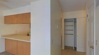 This single-floor space is designed with functionality and practicality in mind. The layout includes a kitchen area visible through a pass-through opening framed with warm wood-toned cabinetry, providing a convenient connection between the cooking and living areas. The cabinetry features a natural wood finish that adds warmth to the otherwise neutral color palette of white and beige tones found throughout.

A notable feature of the space is the built-in shelving unit, which offers multiple tiers of open storage, ideal for organizing books, décor, or everyday essentials. Adjacent to the shelving, a closet with a bi-fold door provides additional concealed storage with a hanging rod and shelves visible within.

The flooring transitions between what appears to be tile in the main area, contributing to a clean, easy-to-maintain surface. The textured ceiling adds subtle visual depth, while the white walls keep the space feeling bright and neutral. A wall-mounted control panel is also present, likely serving as a thermostat or intercom unit. Overall, the space presents a straightforward, well-organized layout with ample storage solutions.