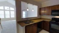 This two-floor space offers a thoughtfully designed interior with a kitchen that serves as a central highlight. The kitchen features rich, dark-stained wood cabinetry paired with warm granite countertops, providing both functionality and visual appeal. A double stainless steel sink sits beneath a large pass-through opening in the wall, which frames a view into the adjacent living area and allows natural light to flow between the spaces. Two pendant lights hang from the ceiling above the counter, adding focused task lighting with a modern touch. Crown molding along the ceiling contributes a refined architectural detail throughout. The kitchen also includes a black dishwasher integrated seamlessly beneath the counter. The pass-through opening reveals an arched architectural detail on the living area side, adding a classic design element. The adjacent living space appears to benefit from generous natural light through large windows. The overall color palette of taupe walls, dark wood tones, and stone-finish counters creates a warm, cohesive ambiance throughout the space. The two-floor layout further enhances the sense of scale and versatility this property offers.