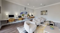 This single-floor space functions as a well-appointed business center and social lounge, thoughtfully designed to accommodate both productivity and relaxation. The interior is finished with warm wood-look flooring throughout, crown molding, and soft neutral gray walls that create a clean, professional atmosphere.

The workstation area features a long, continuous floating desk running along one wall, lined with desktop computers and complemented by woven amber-toned chairs. Potted ornamental grasses and table lamps add warmth to the workspace, while a large abstract painting anchors the wall above the desk. Natural light filters in through black-framed glass doors that open to a lush outdoor courtyard, further brightening the area.

Adjacent to the workstation is a comfortable lounge zone furnished with plush, curved cream armchairs arranged around a patterned area rug. Multiple wall-mounted flat-screen televisions provide entertainment, and curated artwork — including a colorful world map canvas — decorates the walls. Glass-paned French doors connect to an additional room visible in the background, enhancing the sense of openness. Recessed lighting throughout ensures consistent illumination across the entire space.