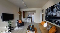 This modern single-floor residence offers a thoughtfully designed open-concept layout that seamlessly connects the living, dining, and kitchen areas. The living room is anchored by a sleek dark leather armchair with vibrant orange accent pillows and a comfortable tan sofa, centered around a contemporary coffee table atop an orange area rug. Track lighting runs along the ceiling, providing ample illumination throughout the space, complemented by natural light that brightens the hardwood-style flooring.

The kitchen is fully equipped with stainless steel appliances, including a full-size refrigerator and microwave, set against warm wood cabinetry. A kitchen island with bar-height seating provides a casual dining option and serves as a natural divider between the cooking and living spaces. Recessed lighting in the kitchen adds a warm, functional glow.

The neutral, warm-toned walls throughout create a cohesive and inviting ambiance, while decorative wall art adds a touch of personality. A hallway visible from the main living area suggests additional private rooms beyond, contributing to a well-organized and functional floor plan. The overall design balances practicality with modern aesthetic appeal, making it a comfortable and stylish living space.