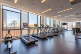 This contemporary fitness center is designed to deliver a comprehensive workout experience in a bright, professionally appointed setting. The space is laid out across a single floor in an angular configuration, allowing for distinct workout zones while maintaining an open, airy feel throughout.

Floor-to-ceiling windows line the perimeter, flooding the interior with natural light and offering sweeping views of the surrounding cityscape and waterfront. The blue rubberized flooring provides both comfort and durability underfoot, complementing the clean, modern aesthetic defined by a drop-panel ceiling with recessed and linear LED lighting.

The cardio zone is anchored by a row of treadmills, elliptical machines, and stationary bikes, all positioned to take full advantage of the exterior views. Flat-screen TVs are mounted throughout for added entertainment during workouts. The strength training area features a multi-functional cable machine, a comprehensive dumbbell rack ranging across multiple weight increments, adjustable benches, and additional free weight equipment. A dedicated functional fitness area includes stability balls, balance tools, and open floor space for stretching or bodyweight exercises.

Textured stone accent walls add visual interest, while a built-in water fountain and bottle-filling station provide practical convenience. The space is well-ventilated and thoughtfully organized to accommodate a wide range of fitness activities.