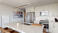 This thoughtfully designed single-floor residence blends contemporary architecture with an abundance of natural light, creating a bright and welcoming atmosphere throughout. Light hardwood flooring flows seamlessly from room to room, complementing the clean, neutral palette of white walls and modern trim.

The kitchen serves as a functional centerpiece, featuring an expansive quartz-topped island with seating for multiple guests, white shaker cabinetry extending to the ceiling, stainless steel appliances, subway tile backsplash, and a sleek linear pendant light overhead. The open layout connects the kitchen directly to the living area, where floor-to-ceiling windows and exposed concrete columns frame sweeping views of the surrounding cityscape and waterfront beyond.

The living space is generously proportioned, bathed in natural light from large multi-panel windows that provide panoramic water views — a standout feature of the residence. The bedroom continues this theme, with a dedicated window offering direct, unobstructed views of the waterfront, creating a tranquil and scenic retreat.

The bathroom is finished in a clean, all-white palette with a full soaking tub and shower surround. A crisp hallway with recessed lighting connects the private areas of the home, maintaining the cohesive, modern aesthetic found throughout the entire space.