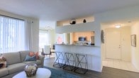 This single-floor residence showcases a thoughtfully designed open-concept layout that effortlessly connects the main living spaces. The kitchen is a functional centerpiece, equipped with stainless steel appliances including a refrigerator and microwave, warm cabinetry, and a raised breakfast bar with a granite-style countertop. Three sleek, modern bar stools line the bar, making it an ideal spot for casual dining or entertaining. The kitchen opens directly to the dining area, which features a dining table set accented with coral and blue tones, positioned beside a large window that floods the space with natural light. The living area is warmly appointed with a plush sofa, a soft area rug in muted tones, and contemporary décor that adds personality without overwhelming the space. Hardwood-style flooring flows throughout, tying the areas together cohesively. The light, neutral white walls and textured ceiling enhance the airy feel of the interior. Decorative accents, including wall art and curated shelf displays above the kitchen pass-through, add character and charm. The overall ambiance is bright, welcoming, and move-in ready, offering a comfortable and stylish environment for everyday living.