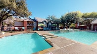 This outdoor pool area offers a well-appointed recreational retreat surrounded by mature shade trees and the classic brick architecture of the surrounding residential buildings. The centerpiece is a generously sized swimming pool with crystal-clear water, accessible via built-in entry steps and metal handrails, complemented by a raised spa featuring decorative stone coping and fountain jets. A broad concrete sundeck surrounds the pool on multiple sides, providing ample space for lounging in the numerous poolside chaise lounges arranged throughout the area. Shaded outdoor seating is available beneath vibrant green market umbrellas alongside tables and chairs, ideal for relaxing out of the sun. The pool area is enclosed by a black metal perimeter fence with a gated entrance, ensuring a secure environment. A low-profile brick clubhouse building runs along one side of the deck, offering covered access to restrooms, a change room, and vending machines for added convenience. The entire space enjoys abundant natural sunlight under open Texas skies, with mature oak trees providing natural shade and enhancing the overall ambiance of this inviting community outdoor space.