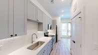 This efficiently designed studio offers a thoughtful single-floor layout that maximizes every square foot. The galley-style kitchen is a standout feature, showcasing floor-to-ceiling gray shaker cabinetry with matte black hardware, crisp white quartz countertops, a hexagonal tile backsplash, and a full suite of stainless steel appliances including a refrigerator, electric range, and built-in microwave. Recessed lighting illuminates the space, while under-cabinet lighting adds a refined touch. Light wood-look flooring runs consistently throughout the unit, creating visual continuity and a warm, cohesive aesthetic. The open living area is bright and airy, benefiting from natural light filtering through a full-length glass exterior door that leads outside, adding both openness and accessibility. The bathroom is clean and functional, featuring a tub-shower combination with white tile surround, brushed nickel fixtures, a white quartz vanity, gray shaker storage cabinetry, and matching wood-look flooring. The overall design palette—neutral whites, soft grays, and warm wood tones—gives the studio a modern yet inviting character that makes efficient use of its compact footprint.