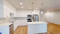 This well-designed 2-bedroom, 1-bathroom residence offers a thoughtfully arranged single-floor layout that maximizes both functionality and comfort. The open-concept living and kitchen area serves as the heart of the home, featuring light-toned hardwood flooring throughout that adds warmth and visual continuity. Large windows allow natural light to fill the living space, with views of the surrounding building exteriors and balconies.

The kitchen is a standout feature, showcasing crisp white cabinetry with sleek hardware, a light gray subway tile backsplash, and a full suite of stainless steel appliances including a range, microwave, and refrigerator. A central island with a white countertop provides additional prep space and is illuminated by two modern pendant lights overhead, creating an inviting focal point.

The bedrooms are carpeted for comfort, with the primary bedroom offering a generous footprint and access to a dedicated walk-in closet equipped with wire shelving systems for organized storage. A dedicated laundry closet houses a full-size side-by-side Whirlpool washer and dryer with additional shelving above. The unit also includes a wall-mounted PTAC climate control unit in the living area, providing convenient heating and cooling throughout the space.