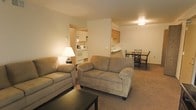 This single-floor space is designed with an open-concept layout that seamlessly connects the living room, dining area, and kitchen. The living room serves as the central gathering space, furnished with two plush sofas in a warm taupe tone, complemented by carpeted flooring and a low-profile coffee table. Warm ambient lighting from a table lamp adds a cozy atmosphere to the space.

A partial dividing wall separates the living area from the kitchen without fully closing off the space, maintaining an open and airy feel. The kitchen is compact yet functional, featuring white cabinetry, a white range oven, a microwave, and a raised breakfast bar with counter seating. Light wood upper cabinets add a subtle contrast to the neutral color palette.

Adjacent to the kitchen, a dining area accommodates a small table with dark wood chairs, providing a dedicated space for meals. The overall design is practical and cohesive, with neutral walls, consistent carpeting in the main living zones, and warm overhead lighting throughout. The layout makes efficient use of the available space, creating a comfortable and livable environment.