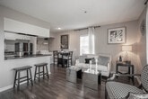 Located at 520 Richey St in Corona, CA, this well-appointed single-story residence offers a thoughtfully designed open-concept layout that seamlessly connects the kitchen, dining, and living areas. Rich wood-look flooring runs throughout the main living spaces, complementing a neutral color palette that lends the interior a clean, contemporary feel.

The kitchen is equipped with white cabinetry, dark granite countertops, black appliances including a refrigerator and range, and a peninsula with bar seating — ideal for casual dining or entertaining. Adjacent to the kitchen, a dedicated laundry area houses a full-size washer and dryer with built-in shelving for added storage convenience.

The living area is bright and welcoming, with natural light filtering through sheer curtains framing a window with outdoor views. A mirrored coffee table and stylish accent chairs contribute to the room's polished aesthetic.

The bedroom is a calm retreat, featuring soft carpeting, a ceiling fan, and a window dressed with sheer panels that allow diffused natural light. The full bathroom includes a tub-shower combination with tile surround, granite countertop vanity, and wood-look flooring, maintaining the cohesive design found throughout the space.