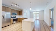 This well-appointed single-floor residence offers a thoughtfully designed open-concept layout that seamlessly connects the kitchen, dining, and living areas. The kitchen is a standout feature, equipped with warm wood-tone cabinetry, stainless steel appliances including a refrigerator with ice maker, electric range, and microwave, along with a large white-topped island and pendant lighting overhead. Track lighting and a window above the sink provide ample illumination. The living area extends naturally from the kitchen, featuring light hardwood flooring, a ceiling fan, and French doors that invite natural light and outdoor views. A secondary room with matching hardwood floors and French door access offers additional flexible living space. The bedroom is carpeted and brightened by a trio of windows fitted with blinds and a ceiling fan above. The primary bathroom includes a glass-enclosed walk-in shower and tile flooring, while the secondary bathroom features a dark wood vanity with a white countertop and clean, minimalist finishes. A dedicated laundry area with a front-loading washer and hardwood flooring adds everyday convenience. Throughout the residence, crisp white walls, consistent flooring choices, and modern fixtures contribute to a cohesive, contemporary aesthetic.