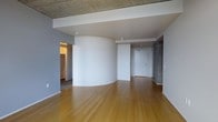 This single-floor space showcases a thoughtful blend of industrial and contemporary design elements. The open main living area immediately draws attention with its distinctive curved white accent wall, which serves as a striking architectural focal point while elegantly dividing the space. Overhead, an exposed concrete ceiling adds raw, industrial character and contrasts beautifully with the smooth white walls and warm-toned hardwood floors that run throughout the unit. The ceiling features a recessed drop section with clean white drywall, creating a layered visual effect that adds depth to the room. A concrete accent panel near the entryway further reinforces the industrial aesthetic. The entryway corridor leads to a solid white door, providing a clean and functional point of entry. To the side, an opening reveals an additional room or walk-in closet area, offering practical storage or living flexibility. The overall layout feels open and airy, with a neutral color palette of whites and grays complemented by the natural warmth of the hardwood flooring. The combination of curved forms, exposed materials, and modern finishes gives this space a distinctive character that balances both functional living and architectural interest.