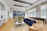 This well-appointed single-floor amenity space is designed to encourage gathering and relaxation, blending contemporary style with warm, inviting details throughout. The main lounge area commands attention with soaring ceilings accented by striking exposed dark wood beams and recessed lighting, complemented by an elegant chandelier. Light hardwood flooring runs continuously throughout, tying the various zones together seamlessly.

The central lounge features a curated mix of seating, including a rich navy velvet sofa, tan armchairs, and a marble-top coffee table, arranged around a soft area rug. Adjacent to this is a generous long-table dining area with a solid wood communal table and upholstered chairs, ideal for group gatherings. A shuffleboard table adds a recreational element nearby.

The wet bar area serves as a focal point, featuring a dramatic floor-to-ceiling blue sculptural tile backsplash, open shelving, white cabinetry, quartz countertops, and a stainless steel sink. A dedicated media lounge offers built-in gray cabinetry, a wall-mounted television, and comfortable neutral seating. Smaller breakout rooms provide intimate settings with round tables and accent chairs.

Tall multi-pane windows along the perimeter flood the space with natural light, enhancing the open, airy atmosphere throughout.