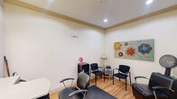 Located at 31511 Nichols Sawmill Road in Magnolia, Texas, this single-room salon suite offers a fully functional layout designed to accommodate professional hair care services. The space is finished with warm wood-look flooring throughout, neutral cream-toned walls, and crown molding details that lend a clean, polished appearance.

The room is arranged to serve multiple functions simultaneously. A dedicated styling station features a wall-mounted mirror with overhead vanity lighting, a built-in shampoo bowl with an integrated backwash unit, and a professional styling chair positioned to make efficient use of the available floor area. An additional styling chair with a hooded dryer occupies a separate corner, expanding the service capacity of the suite.

A seating area along one wall provides a comfortable waiting space, furnished with padded guest chairs arranged around a small side table with a lamp. A large, colorful floral canvas adds a vibrant decorative focal point to the room. Recessed and surface-mounted lighting ensures the space is well-lit throughout. A frosted glass door with a private entry leads into the suite, and access to an adjacent common area is visible through a secondary doorway, connecting the suite to the broader facility.