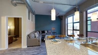 This stylish single-floor space showcases a seamless open-concept layout that blends industrial design with contemporary comfort. Exposed ductwork runs along the high ceilings, adding a loft-like character that complements the clean, modern aesthetic throughout. The living area is anchored by a bold teal accent wall, which creates a striking focal point and adds depth to the space. A generous sectional sofa, paired with mid-century modern accent furniture, gives the lounge area a comfortable yet design-forward feel. Large floor-to-ceiling windows flood the interior with natural light and offer views of the surrounding streetscape, enhancing the sense of openness. The dining area flows naturally from the living space, centered around a sleek table with wood and metal chairs that echo the industrial-modern theme. A granite-topped kitchen island serves as a visual and functional divider between the kitchen and the dining area, providing additional surface space for meals or casual gatherings. Pendant lighting with cylindrical shades is suspended above, offering warm, focused illumination. The flooring throughout features wood-look planks that tie the entire open floor plan together cohesively, making this space as practical as it is visually compelling.
