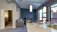 This stylish single-floor space showcases a seamless open-concept layout that blends industrial design with contemporary comfort. Exposed ductwork runs along the high ceilings, adding a loft-like character that complements the clean, modern aesthetic throughout. The living area is anchored by a bold teal accent wall, which creates a striking focal point and adds depth to the space. A generous sectional sofa, paired with mid-century modern accent furniture, gives the lounge area a comfortable yet design-forward feel. Large floor-to-ceiling windows flood the interior with natural light and offer views of the surrounding streetscape, enhancing the sense of openness. The dining area flows naturally from the living space, centered around a sleek table with wood and metal chairs that echo the industrial-modern theme. A granite-topped kitchen island serves as a visual and functional divider between the kitchen and the dining area, providing additional surface space for meals or casual gatherings. Pendant lighting with cylindrical shades is suspended above, offering warm, focused illumination. The flooring throughout features wood-look planks that tie the entire open floor plan together cohesively, making this space as practical as it is visually compelling.