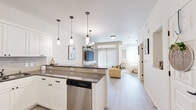 This thoughtfully designed single-floor unit showcases a clean, contemporary aesthetic defined by bright white walls, light hardwood-style flooring, and a cohesive neutral palette accented with warm gold tones. The open-concept layout seamlessly connects the kitchen, dining, and living areas, creating an airy and functional flow throughout the space. The kitchen is a standout feature, equipped with full stainless steel appliances including a French door refrigerator, electric range, over-the-range microwave, and dishwasher. Crisp white shaker-style cabinetry extends to the ceiling, complemented by a grey tile backsplash and dark countertops. A peninsula with bar seating and elegant pendant lighting doubles as a casual dining and entertaining space, while a separate dining area accommodates a full table. The living area benefits from a large sliding glass door dressed with vertical blinds, filling the room with natural light. A built-in media wall adds a modern focal point. The bedroom is generously proportioned, featuring sliding glass door access, large windows, and ample natural light. The bathroom is finished with a white vanity, dark countertop, a framed mirror, and stylish light fixture, completing the unit's refined and modern character.