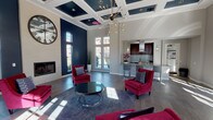 This striking single-floor communal space exudes contemporary sophistication through a carefully curated blend of bold design choices and refined finishes. The centerpiece of the room is its dramatic, soaring coffered ceiling, painted in deep navy blue and accented with recessed lighting, which draws the eye upward and amplifies the grandeur of the space. A sculptural Sputnik-style chandelier hangs prominently, serving as a dazzling focal point that anchors the lounge area below.

The open floor plan is expansive and bathed in natural light, courtesy of numerous large multi-pane windows and glass French doors that line the perimeter, offering outdoor views and a bright, welcoming atmosphere. Bold navy accent walls contrast elegantly against the light gray walls and white trim throughout the space.

The lounge area features plush crimson velvet armchairs arranged around a sleek glass and chrome coffee table atop a gray area rug, creating an inviting conversation setting. A classic gas fireplace with an oversized decorative wall clock above it adds warmth and character to the room.

Adjacent to the lounge, a well-appointed kitchenette showcases rich mahogany cabinetry, a mosaic tile backsplash, quartz countertops, a stainless steel sink, and bar seating — ideal for entertaining. The flooring transitions seamlessly between large-format tile and wood-look planks, tying the entire space together with a polished, modern aesthetic.