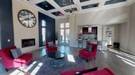 This striking single-floor communal space exudes contemporary sophistication through a carefully curated blend of bold design choices and refined finishes. The centerpiece of the room is its dramatic, soaring coffered ceiling, painted in deep navy blue and accented with recessed lighting, which draws the eye upward and amplifies the grandeur of the space. A sculptural Sputnik-style chandelier hangs prominently, serving as a dazzling focal point that anchors the lounge area below.

The open floor plan is expansive and bathed in natural light, courtesy of numerous large multi-pane windows and glass French doors that line the perimeter, offering outdoor views and a bright, welcoming atmosphere. Bold navy accent walls contrast elegantly against the light gray walls and white trim throughout the space.

The lounge area features plush crimson velvet armchairs arranged around a sleek glass and chrome coffee table atop a gray area rug, creating an inviting conversation setting. A classic gas fireplace with an oversized decorative wall clock above it adds warmth and character to the room.

Adjacent to the lounge, a well-appointed kitchenette showcases rich mahogany cabinetry, a mosaic tile backsplash, quartz countertops, a stainless steel sink, and bar seating — ideal for entertaining. The flooring transitions seamlessly between large-format tile and wood-look planks, tying the entire space together with a polished, modern aesthetic.