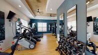 This fitness center offers a thoughtfully designed workout environment that balances aesthetics with functionality. The space features striking teal accent walls paired with crisp white trim, creating a vibrant and energizing atmosphere. Light hardwood flooring runs throughout, providing a clean, durable surface suitable for a variety of exercises.

The ceiling is a standout architectural detail, showcasing a coffered tray design with warm wood paneling and dual ceiling fans that ensure comfortable airflow during workouts. Recessed lighting supplements the abundant natural light that streams in through multiple windows fitted with white blinds.

The fitness floor is comprehensively equipped for both cardio and strength training. Cardio offerings include treadmills, elliptical machines, a StairMaster, and a stationary spin bike, each paired with wall-mounted flat-screen televisions for entertainment. A multi-station cable machine and a heavy bag provide versatile strength and conditioning options. A full dumbbell rack with floor-to-ceiling mirrors lines one wall, making it easy to monitor form during free weight training.

Near the entrance, a welcoming area includes yoga mat storage, a mini-fridge, and a water dispenser for added convenience. A restroom is accessible directly from the fitness floor. The glass entry door opens to an outdoor area, allowing natural light to flow in and giving the space an open, inviting feel.