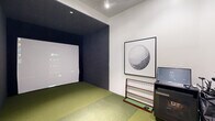 Located at 3955 Design Center Dr, this dedicated indoor golf simulator room offers a fully equipped, self-contained environment designed for serious practice and recreational play. The space features a lush artificial turf floor that spans the entire room, creating an authentic course-like feel underfoot. A large dark-walled hitting bay anchors one end of the room, housing a full-sized projection screen that displays simulation software, delivering an immersive virtual golf experience. The room is powered by a TrackMan launch monitor system, a leading technology in shot tracking and performance analysis, supported by a dedicated workstation and club rack for convenient equipment access. A ceiling-mounted projector delivers crisp visuals onto the screen, while recessed lighting illuminates the surrounding space cleanly. The white walls of the main area contrast with the deep, dark interior of the hitting bay, clearly delineating the playing zone. A glass-paneled entry door allows for easy access, and golf simulator rules are posted near the entrance for user guidance. Decorative framed golf ball artwork adorns the wall, adding a polished, thematic touch to the overall design. The room is compact yet highly functional, providing everything needed for a comprehensive indoor golf session.