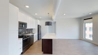 This single-floor, 2-bedroom, 2-bathroom residence offers a thoughtfully designed open-concept layout that seamlessly connects the kitchen and living areas. The contemporary kitchen is a standout feature, appointed with stainless steel appliances including a refrigerator, range, and over-the-range microwave. Crisp white cabinetry extends to the ceiling, maximizing storage, while a gray tile backsplash adds texture and visual interest. A dark-base kitchen island topped with a light quartz-style countertop serves as a functional centerpiece, complemented by a stylish cylindrical drum pendant light overhead. Recessed lighting is integrated throughout the ceiling, ensuring consistent, bright illumination across the open space. Light wood-toned flooring runs throughout, contributing to a clean, modern aesthetic and a sense of cohesion between areas. The living space beyond the kitchen is open and airy, offering a versatile canvas for various furniture arrangements. The overall ambiance is fresh and contemporary, with a neutral color palette of whites and soft grays that lends the interior a polished, move-in-ready appeal suitable for a range of lifestyles.