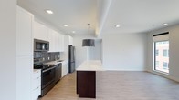This single-floor, 2-bedroom, 2-bathroom residence offers a thoughtfully designed open-concept layout that seamlessly connects the kitchen and living areas. The contemporary kitchen is a standout feature, appointed with stainless steel appliances including a refrigerator, range, and over-the-range microwave. Crisp white cabinetry extends to the ceiling, maximizing storage, while a gray tile backsplash adds texture and visual interest. A dark-base kitchen island topped with a light quartz-style countertop serves as a functional centerpiece, complemented by a stylish cylindrical drum pendant light overhead. Recessed lighting is integrated throughout the ceiling, ensuring consistent, bright illumination across the open space. Light wood-toned flooring runs throughout, contributing to a clean, modern aesthetic and a sense of cohesion between areas. The living space beyond the kitchen is open and airy, offering a versatile canvas for various furniture arrangements. The overall ambiance is fresh and contemporary, with a neutral color palette of whites and soft grays that lends the interior a polished, move-in-ready appeal suitable for a range of lifestyles.