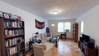 This single-floor space showcases a welcoming living room as its central feature. The room is finished with classic parquet hardwood flooring, laid in a distinctive geometric pattern that adds visual warmth and character to the space. Neutral white walls keep the atmosphere light and versatile, complementing the flush ceiling light fixture that provides overhead illumination.

Natural light enters through a front-facing entrance door with a fan-shaped transom window, alongside an adjacent window fitted with a window air conditioning unit, ensuring year-round comfort. The layout accommodates a sectional sofa and upholstered armchair arranged around a rectangular coffee table, creating a practical and inviting seating area.

A baseboard heating unit runs along the far wall, indicating a reliable in-unit heating system. Built-in or freestanding bookshelves flank the room, offering ample storage and display space for books and personal items. A decorative textile wall hanging adds a personalized artistic touch to the otherwise understated décor. The overall ambiance is calm and functional, with a straightforward layout that maximizes the usable living area.