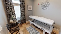 This single-floor wellness space is designed to offer a refined and comfortable experience for guests. The tanning suite is the centerpiece of the space, featuring a professional-grade SunMaster tanning bed positioned within a neatly appointed room that balances function with aesthetic appeal.

The room is finished with soft gray walls that create a calm, neutral backdrop, complemented by natural stone tile flooring that adds texture and warmth underfoot. A decorative gray striped accent rug is placed in front of the tanning bed, adding a cozy touch to the space. Bold black-and-white patterned curtains frame a bright window, allowing natural light to filter in while maintaining privacy and adding a striking visual contrast to the subdued wall tones.

A small bistro-style table and chair set is tucked beside the window, providing a relaxing spot before or after a session. Wall décor includes a framed architectural print and an intricate geometric metal wall accent, contributing an elevated, boutique-salon feel to the room.

A fire alarm is visible on the wall, indicating the space adheres to safety standards. Overall, the suite strikes a balance between practicality and style, offering guests a welcoming, well-equipped environment for their tanning experience.