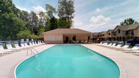 Located at 1655 Gilmore Park Ave in Columbus, Ohio, this single-story community amenity space is designed to offer residents a welcoming and functional outdoor retreat. The centerpiece of the property is a generously sized outdoor swimming pool with crystal-clear turquoise water, framed by a wide concrete deck that provides ample room for relaxation and recreation. Rows of lounge chairs line both sides of the pool, offering comfortable spaces to unwind in the sun or shade. A covered patio area adjacent to the pool house features outdoor seating with tables and chairs, making it well-suited for casual gatherings. The pool house itself is a compact, single-story structure with a neutral stucco and brick exterior topped by a brown hip roof. It includes an open-air passage that connects the pool deck to the surrounding grounds, enhancing the flow of the outdoor space. The area is enclosed by a black iron fence, ensuring safety and privacy. Lush, mature trees rise in the background, providing a natural canopy that adds to the serene ambiance. The overall setting is clean, well-maintained, and thoughtfully arranged to support an active and social community lifestyle.