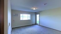 This single-floor space offers a straightforward and functional layout designed for comfortable everyday living. The main living area is the focal point of the space, featuring neutral, light-toned walls and soft carpeted flooring that lend a clean and understated aesthetic throughout.

Natural light is a notable characteristic of this room, provided by a large multi-pane window that frames views of a green, open outdoor area. A glass-panel exterior door further enhances the connection to the outdoors while allowing additional light to fill the space. Together, these openings create a bright and airy atmosphere.

Climate control is well-equipped, with a wall-mounted air conditioning unit installed alongside the window and baseboard heating running along the lower wall — ensuring year-round comfort. A centrally mounted ceiling light fixture provides reliable ambient illumination.

The room's clean lines, simple finishes, and practical features make it a versatile space well-suited for a variety of uses. The open layout, combined with the natural light and outdoor access, gives the space an inviting and livable quality that is both functional and easy to personalize.
