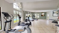 Located at 11247 San Jose Blvd in Jacksonville, FL, this single-floor fitness facility offers a comprehensive workout environment across a generously sized footprint. The exercise room is the centerpiece of the space, divided into distinct functional zones. A bright cardio area features treadmills and stationary bikes positioned beneath a dramatic arched window that floods the room with natural light, while large windows and glass doors frame views of an outdoor pool and lush greenery. The strength training zone is finished with hardwood flooring and rubber matting, and is equipped with cable machines, a dumbbell rack, adjustable benches, medicine balls, and a wall-mounted mirror. A drop ceiling with recessed lighting and ceiling fans ensures a comfortable environment throughout. The walls feature a stylish two-tone striped design in neutral tones, adding a modern, polished aesthetic. A dedicated kitchen complements the facility, outfitted with dark wood cabinetry, granite countertops, stainless steel appliances including a refrigerator, microwave, and range, and a peninsula with bar seating. Light tile flooring runs through the kitchen and entry area, which features soaring ceilings with elegant arched architectural detailing, creating a welcoming transition into the workout space.